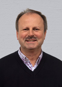 Portrait von Johannes Streitberger, lächelnd mit Schnauzer, kariertem Hemd und schwarzem Pullover obendrüber