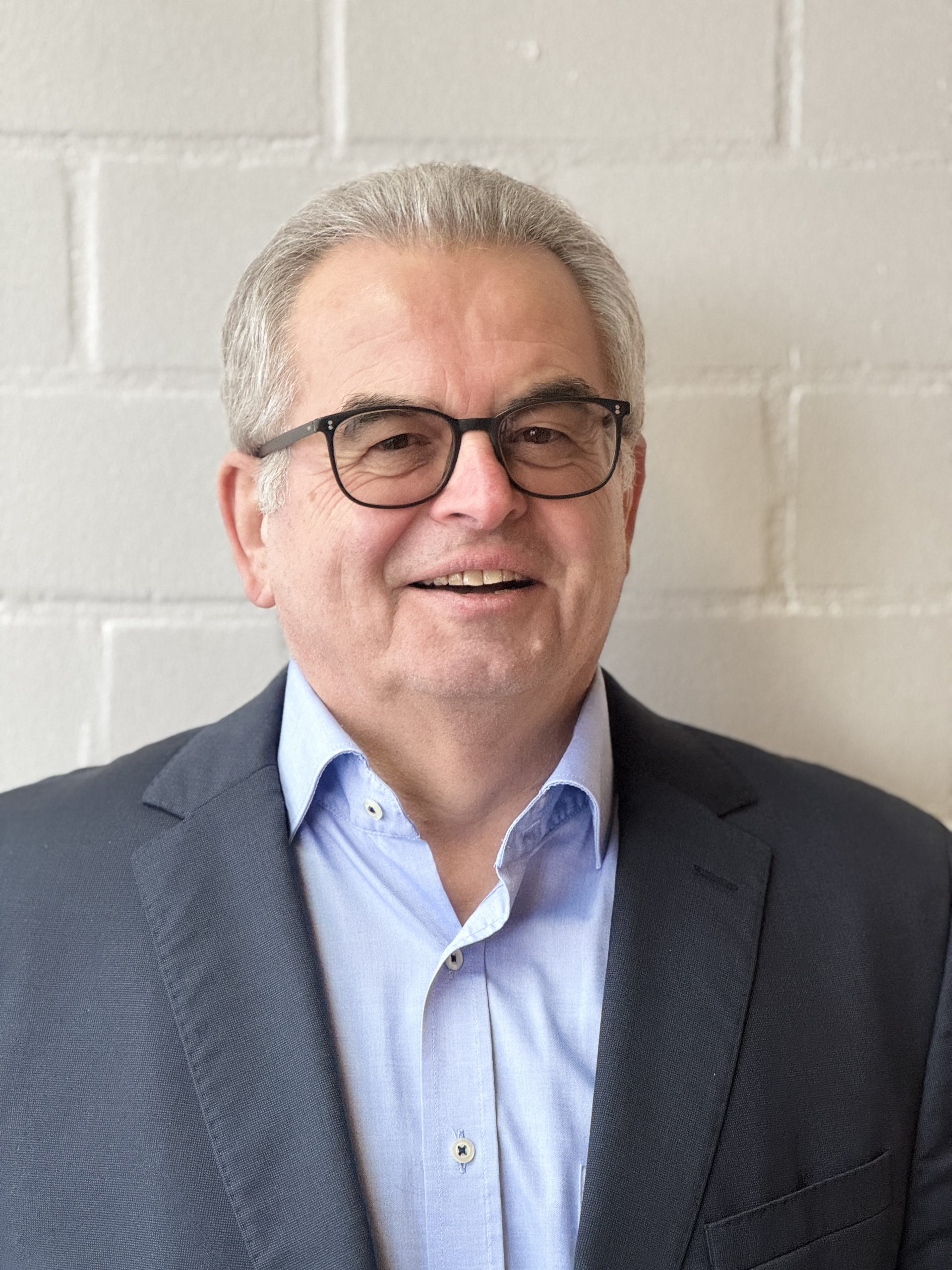 2026 Josef Schneller Das farbige Foto zeigt den Kandidat für den Pfarrgemeinderat St. Korbinian Josef Schneller vor einer weißen Wand, mit Brille, kurzen grauen Haaren, hellblauen Hemd und grauer Anzugsjacke