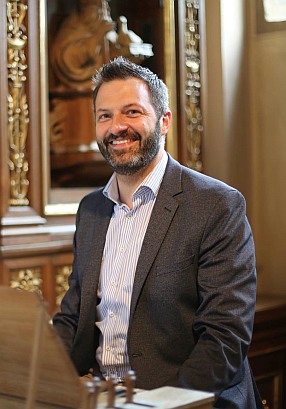 Foto farbig Kirchenmusiker St. Ulrich Foto farbig, Kirchenmusiker St. Ulrich, Matthias Berthel vor gerahmten Bild mit Anzug und weißen Hemd